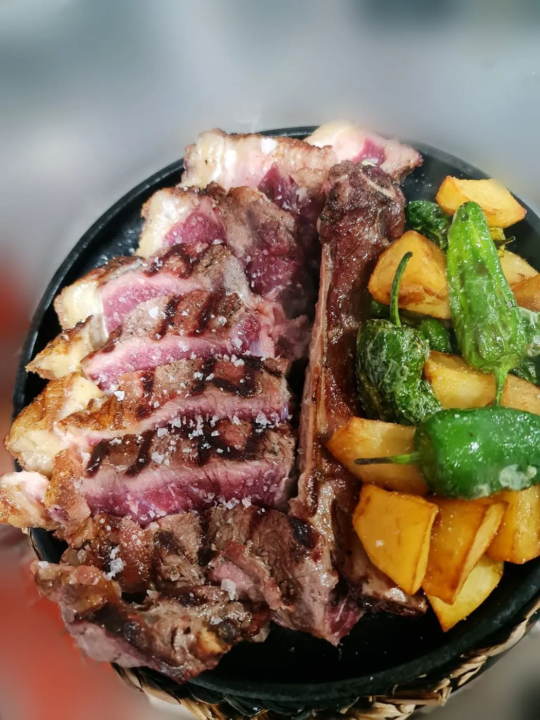 Carne a la brasa cortada en lonchas con patatas y pimientos de Padrón sobre un plato de hierro